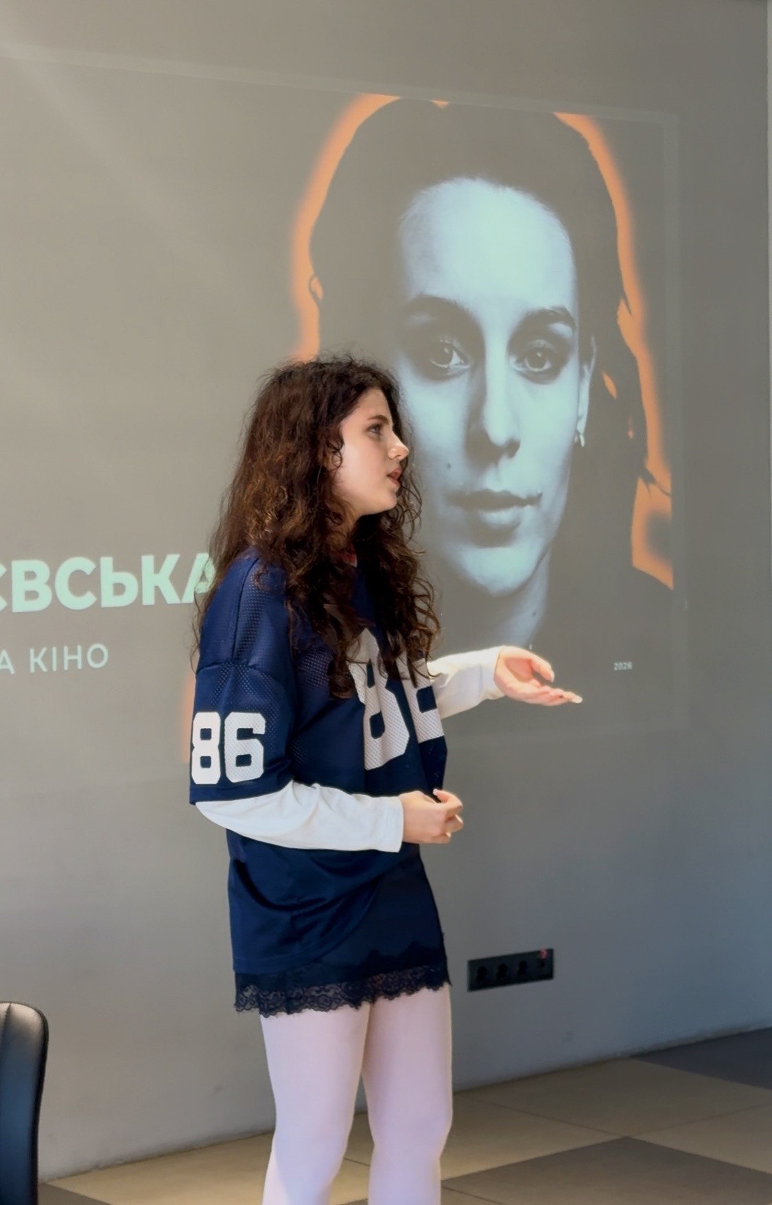 A young woman is standing near a wall, explaining something while gesturing with her hand 